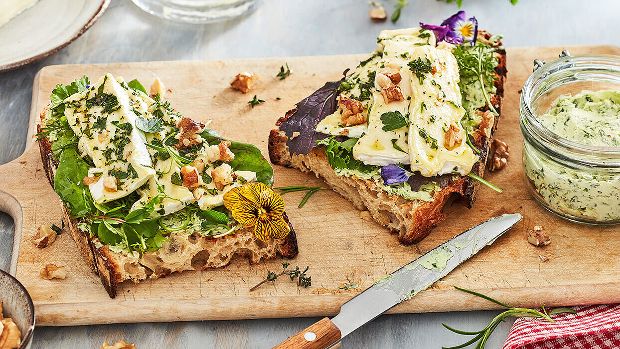 Geröstetes Sauerteigbrot mit mariniertem Camembert und Bärlauch-Butter auf einem Holzbrett garniert mit frischen Kräutern.