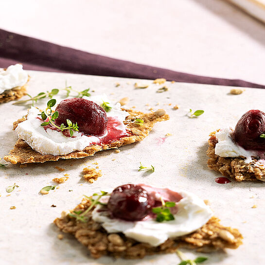 Cracker belegt mit Ziegenkäse und einem Topping aus Ofentrauben, beträufelt mit Honig. Serviert auf einer grauen Schieferplatte