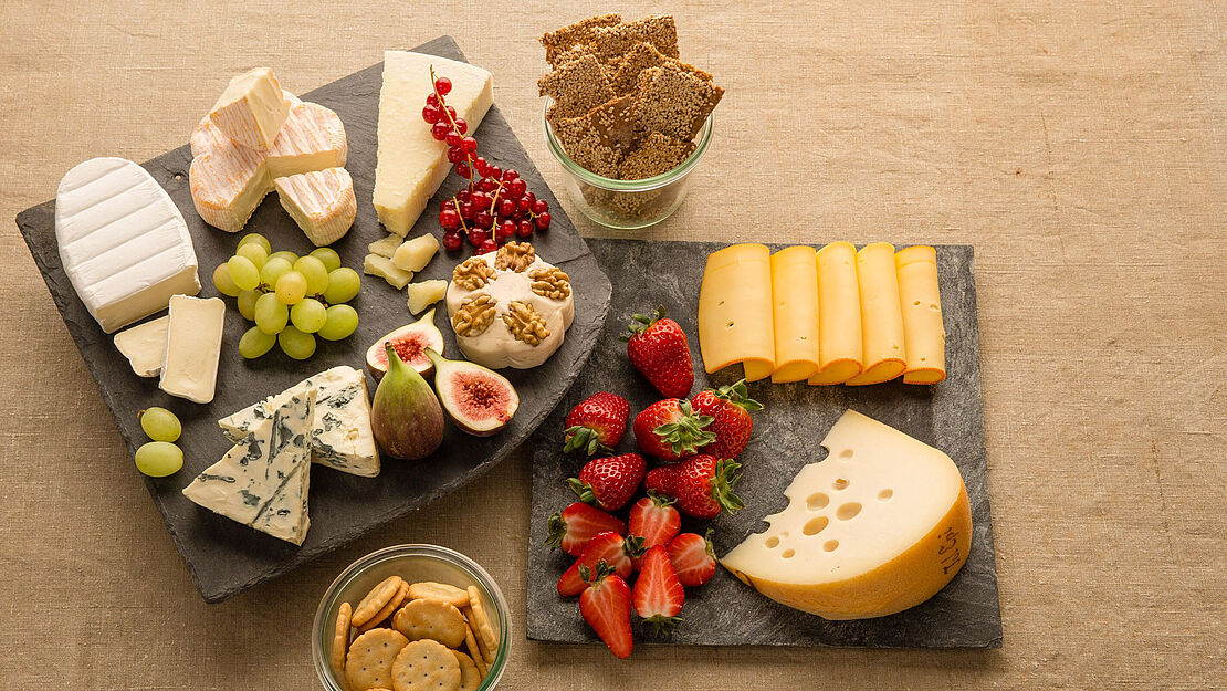 Modische Schieferplatten mit verschiedenen Käsesorten und frischen Obst. Dazu ein paar Cracker im Glas.