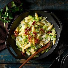 Cremige Schupfnudeln mit zartem Spitzkohl, knusprigem Speck und reichhaltiger Käsesauce – serviert in einer schwarzen Schüssel. 