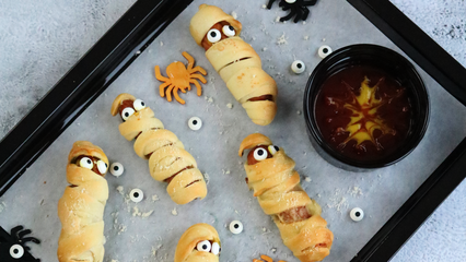 Backblech mit goldbraun gebackenen Würstchen-Mumien aus Pizzateig, verziert mit Zuckeraugen; daneben eine Schale Dip und kleine Halloween-Dekorationen.