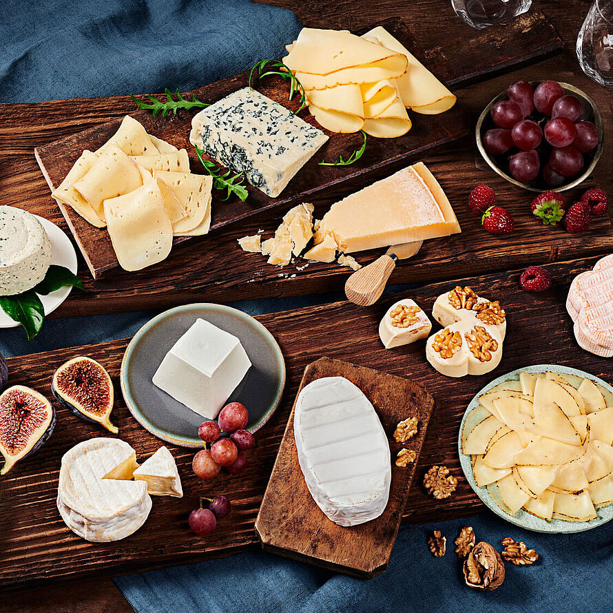 Verschiedene Käsesorten wie BRESSO, Saint Agur und Grana Padano dekorativ auf einer großen Platte angerichtet, begleitet von frischem Obst und Gemüse.