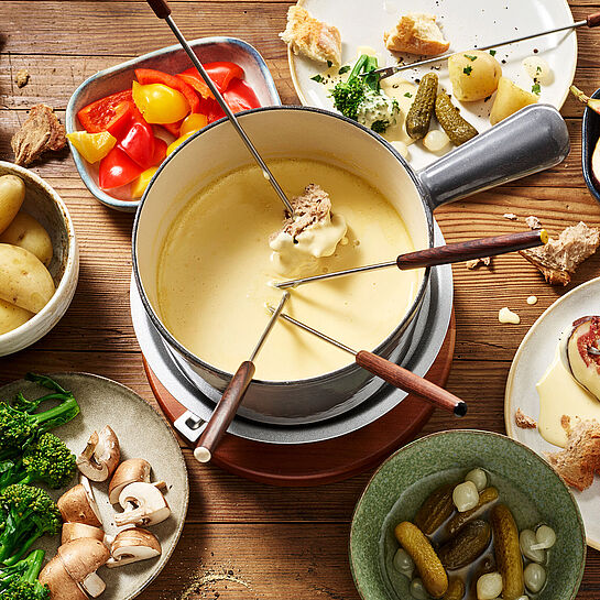 Schön angerichtetes Käsefondue ohne Alkohol mit zahlreichen Zutaten in kleinen Schälchen. Dazu frisches Brot.