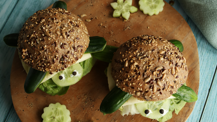 Zwei lustige Schildkrötenbrötchen aus Vollkorn-Saatenbrötchen, Gurken, Käse und Salat, kreativ angerichtet als Pausenbrot für Kinder, auf einem runden Holzbrett serviert.