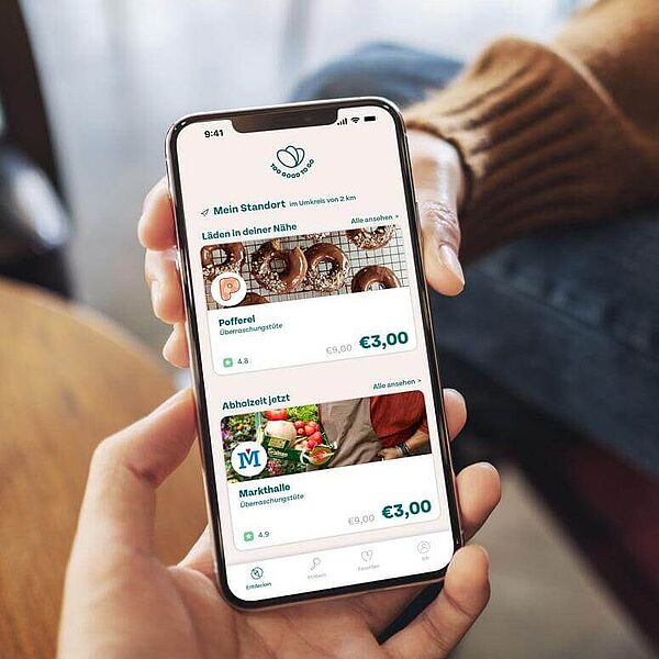 Hand hält ein Handy, dessen Bildschirm die geöffnete App von Too Good To Go zeigt