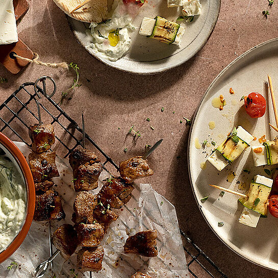 Zweierlei Grillspieße mit Feta und Lammfleisch, serviert mit Tsatsiki auf einem rustikalen Grillrost.