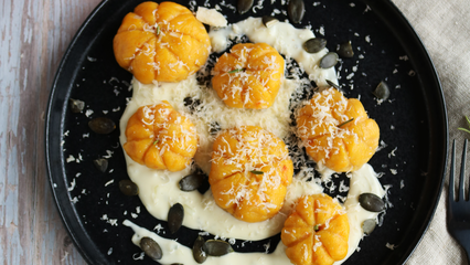 Schwarzer Teller mit kleinen Kürbisgnocchi in Kürbisform, serviert auf cremiger Käsesoße, garniert mit geriebenem Käse und Kürbiskernen.