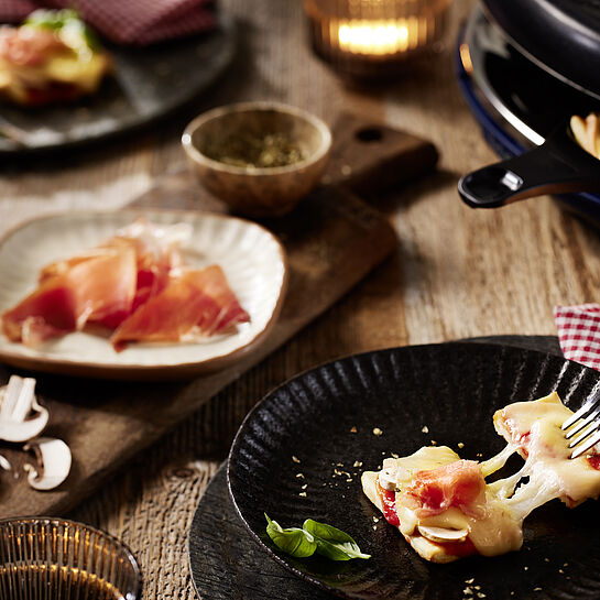 Knuspriger Pizza-Teig trifft auf cremig-würzigen Raclette-Käse von LE RUSTIQUE, gekrönt mit herzhaftem Rohschinken auf einem schwarzen Raclette-Teller