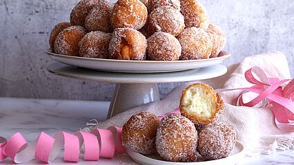 Frisch frittierte Quarkbällchen bestäubt mit feinem Zucker auf einer weißen Kuchenplatte und einem Teller serviert. 