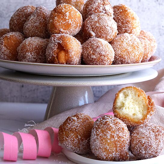 Frisch frittierte Quarkbällchen bestäubt mit feinem Zucker auf einer weißen Kuchenplatte und einem Teller serviert. 