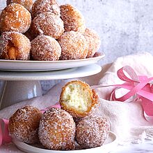 Frisch frittierte Quarkbällchen bestäubt mit feinem Zucker auf einer weißen Kuchenplatte und einem Teller serviert. 