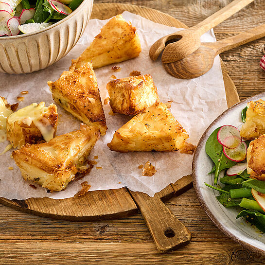 Knusprige Camembert-Ecken aus Filoteig begleitet von einem frischen, grünen Spinatsalat mit Radieschen. Fein angerichtet auf einer rustikalen Holzplatte.