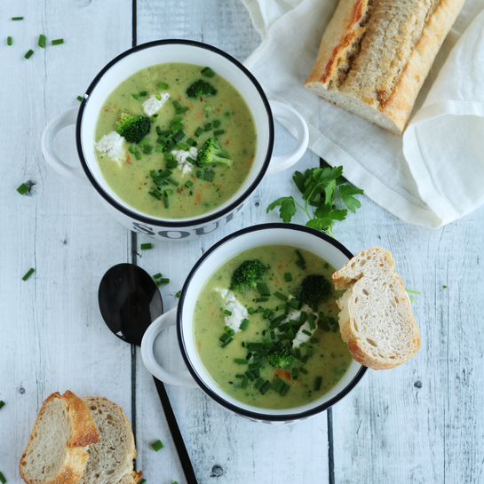 Zwei weiße Suppenschalen mit cremiger Brokkoli-Gemüse-Cremesuppe, garniert mit frischen Kräutern und Baguettescheiben auf einem weißen Holztisch.