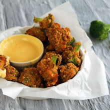 Knusprig panierte, goldbraune Brokkoliröschen werden in einer mit Backpapier ausgelegten Schale serviert, dazu ein Käsedip aus Milkana Gouda; im Hintergrund liegen frische Brokkoliröschen auf einer hellgrauen Oberfläche.