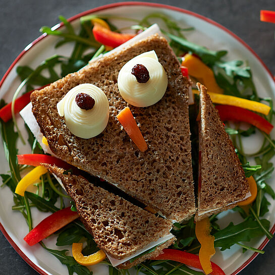 Kindliches Vollkorn-Pausenbrot mit einem drapierten Gesicht aus Käse, Karotten und Rosinen