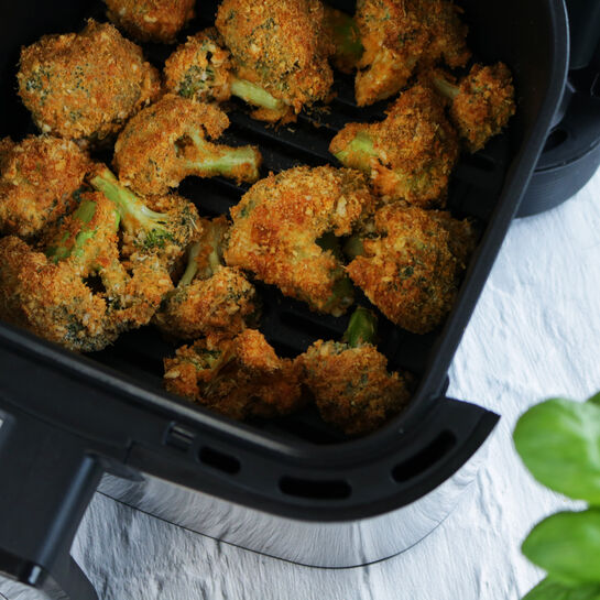 Panierte, goldbraun gebackene Brokkoliröschen liegen in einem schwarzen Airfryer-Korb; im Hintergrund eine helle Oberfläche und ein grünes Basilikumblatt.