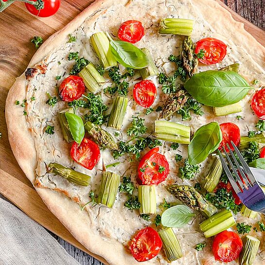 Leckerer Flammkuchen auf einem Holzbrett, belegt mit frischem Spargel, Tomaten, Basilikum und Petersilie