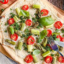 Leckerer Flammkuchen auf einem Holzbrett, belegt mit frischem Spargel, Tomaten, Basilikum und Petersilie