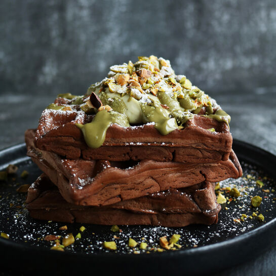 Gestapelte Schoko-Pistazien-Waffeln auf einem schwarzen Teller, verfeinert mit Pistazienstückchen, grünem Pistazien-Creme-Topping, Sahne und Puderzucker.