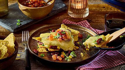 Leckeres Raclettegericht mit knusprigen Tortilla-Chips, geschmolzenem Raclette-Käse, Mais, Bohnen und einer frischen Tomatensalsa, verfeinert mit Koriander.