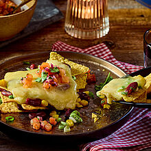 Leckeres Raclettegericht mit knusprigen Tortilla-Chips, geschmolzenem Raclette-Käse, Mais, Bohnen und einer frischen Tomatensalsa, verfeinert mit Koriander.