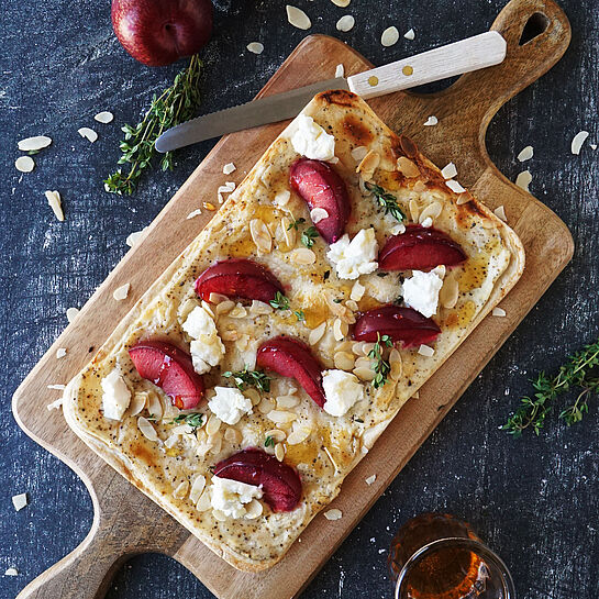 Knuspriger Flammkuchen belegt mit Ziegenfrischkäse, saftigen Pflaumen, Mandeln und einem Hauch Honig, perfekt für Genießer.