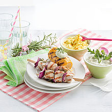 Gegrillte Fleischspieße und ein gegrillter Maiskolben auf einem weißen Teller. Dazu 2 Schüsseln mit Curry Dips. 