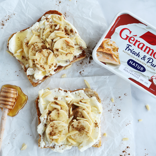 Zwei Scheiben Toast mit Frischkäse, Bananenscheiben, Zimt, Honig und Mandelstiften auf weißem Backpapier. Tolle Toast Idee für eine abwechslungsreiche Brotzeit. 