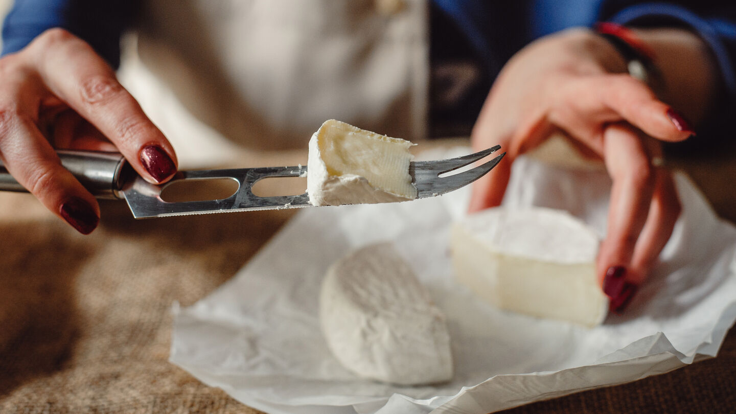 verbraucherpanel-headerbild-2 Ein Stück weicher Weißschimmelkäse wird mit einem Käsemesser über einem ausgepackten Käse auf Pergamentpapier gehalten