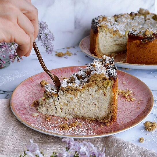 Ein Stück köstlicher Rhabarber-Streuselkuchen mit cremiger Frischkäsefüllung und knusprigen Streuseln auf einem rosa Teller.