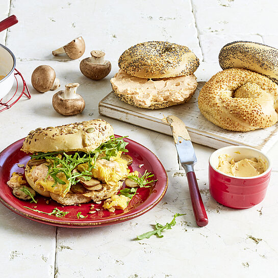 Tolle Frühstückkomposition mit Bagels, Brunch Aufschnitt, Pilzen, Rucola und Rührei. Als Mittelpunkt ein belegtes Frühstückbagel