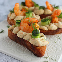 Elegante Canapés aus knusprigem Baguette mit marinierten Schrimps, cremiger Dill-Mousse und frischen Gurkenstreifen, dekorativ angerichtet.