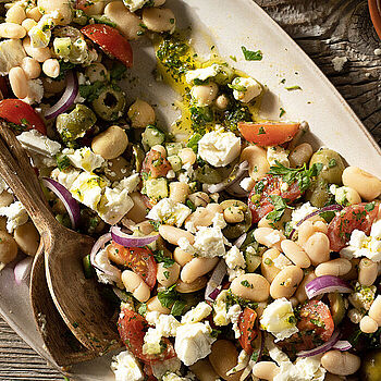 Griechischer Bohnensalat mit Feta, dicken Bohnen, Oliven und frischen Kräutern angerichtet auf einer großen weißen Salatplatte 