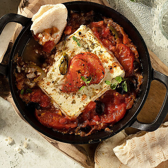 Feta und geschmorte Tomaten frisch aus dem Ofen, serviert in einem Gusseisentopf auf einem Holzbrett, dazu selbstgemachtes Brot.