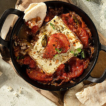 Feta und geschmorte Tomaten frisch aus dem Ofen, serviert in einem Gusseisentopf auf einem Holzbrett, dazu selbstgemachtes Brot.