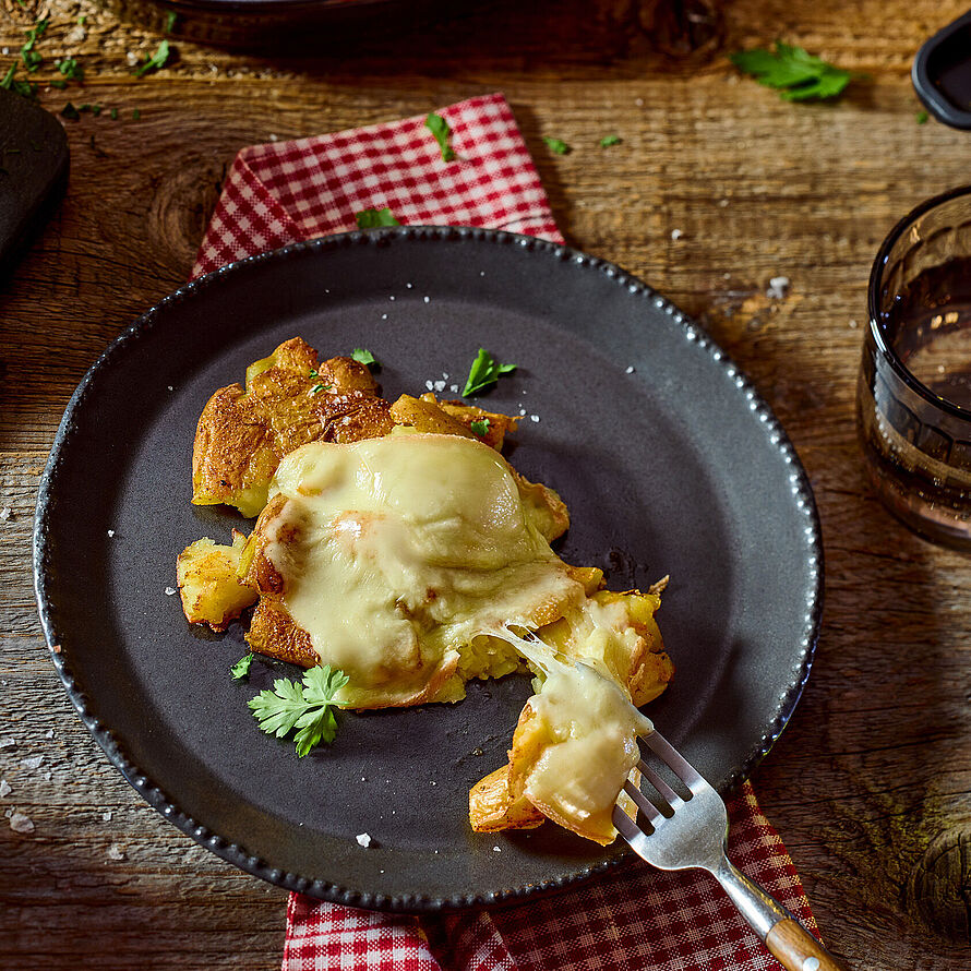 Knusprige, zerdrückte Kartoffeln, goldbraun gegrillt und mit geschmolzenem Raclette-Käse überbacken. Serviert auf einem schwarzen Teller.