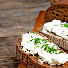 Dunkle Vollkornbrotscheiben mit Frischkäse &amp; Kräutern belegt. Auf einem Holzbrett angerichtet.