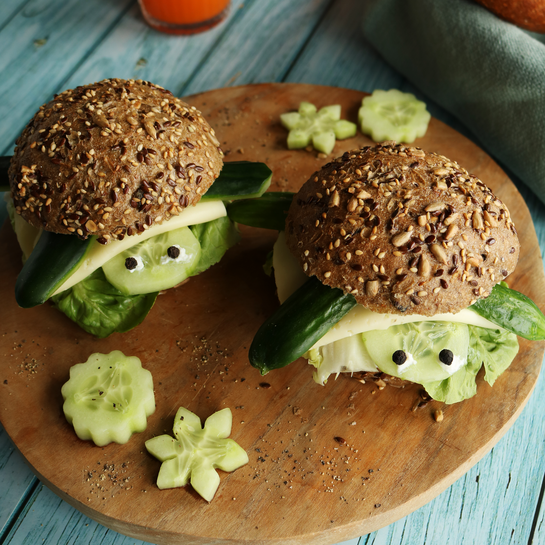 Zwei lustige Schildkrötenbrötchen aus Vollkorn-Saatenbrötchen, Gurken, Käse und Salat, kreativ angerichtet als Pausenbrot für Kinder, auf einem runden Holzbrett serviert.