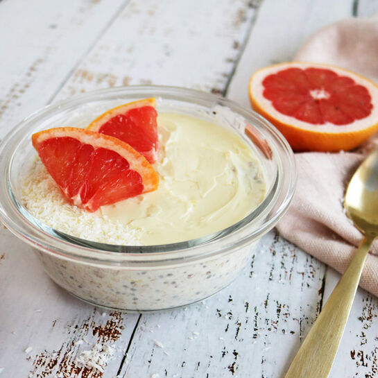 Ein Glas mit cremigen Overnight Oats, garniert mit Grapefruitstücken, steht auf einer weißen Holzoberfläche; eine Hand hält einen Löffel über die Schale, umgeben von halbierten Grapefruits und einem beigefarbenen Tuch.