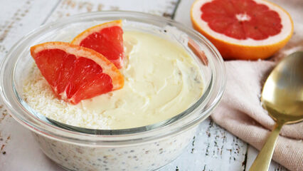 Ein Glas mit cremigen Overnight Oats, garniert mit Grapefruitstücken, steht auf einer weißen Holzoberfläche; eine Hand hält einen Löffel über die Schale, umgeben von halbierten Grapefruits und einem beigefarbenen Tuch.