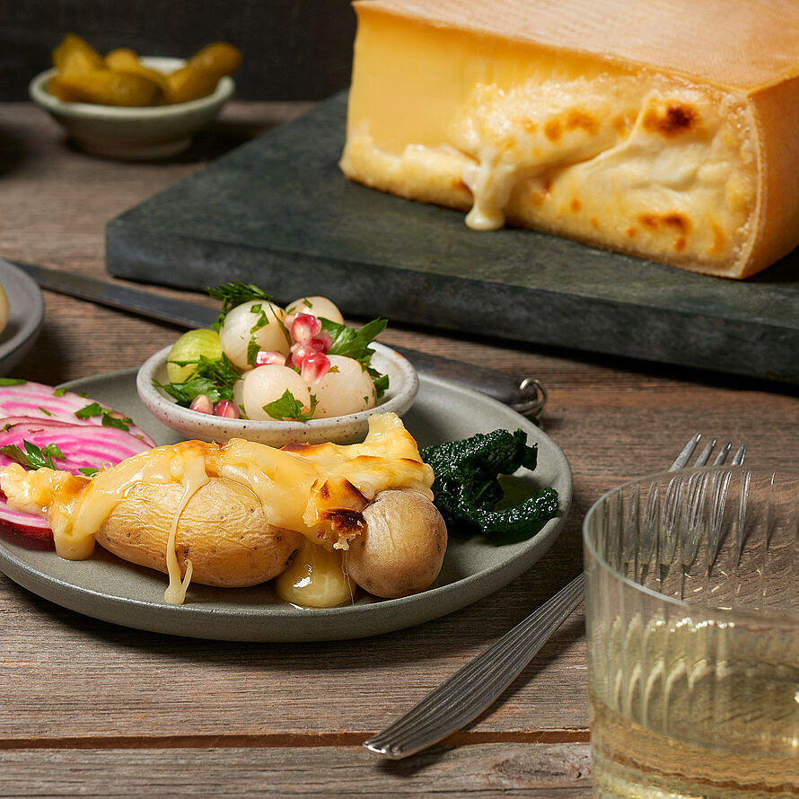 Überbackene Kartoffeln mit Käse und ein Stück Raclette-Käse, serviert auf einer schwarzen Schieferplatte.
