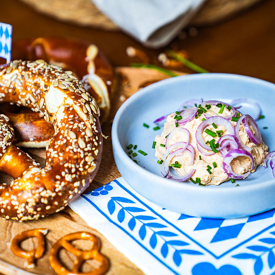 Kleiner bayerischer Snack – Veganer Obazda in einer kleinen blauen Schüssel, serviert mit einer Sesam-Brezel. 