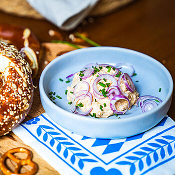 Kleiner bayerischer Snack – Veganer Obazda in einer kleinen blauen Schüssel, serviert mit einer Sesam-Brezel. 