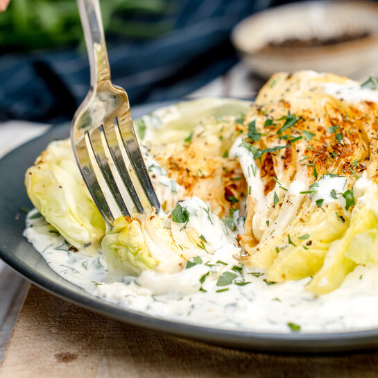 Auf dem Teller liegen geröstete Weißkohlstücke in einer cremigen Kräuter-Käsesoße. Eine Gabel nimmt gerade ein Stück des goldbraun gebratenen Kohls auf. Das Gericht wirkt frisch, würzig und appetitlich angerichtet.