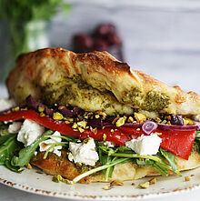 Knuspriges Pizza-Sandwich Panuozzo mit Pesto, Rucola, Feta und Oliven, perfekt für einen schnellen und leckeren Snack – serviert auf einem weißen Teller.