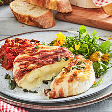 Le Rustique Camembert im Kräuter-Speckmantel, serviert mit Tomatenchutney und Wildkräutersalat. Dazu frisches Baguette und Wasser mit Zitrone. 