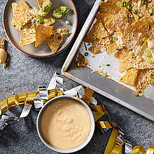 Überbackene Nachos mit Lauch auf einem Backblech. Dazu Käsesauce und Guacamole in kleinen Schüsseln.