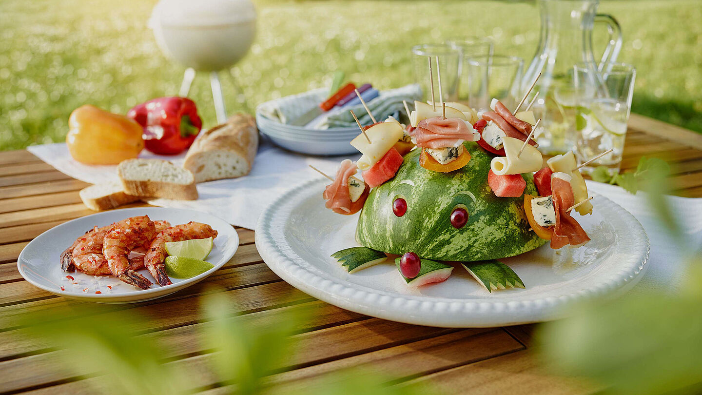 Die exotische Sunny, der Käseigel für den Grillabend Käseigel Sunny mit Saint Agur, Fol Epi, Parmaschinken und Wassermelone bestückt sitzt auf einem Gartentisch