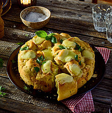 Goldbraunes Raclette-Zupfbrot mit geschmolzenem Käse und Kräutern, angerichtet auf einem rustikalen Holzbrett.