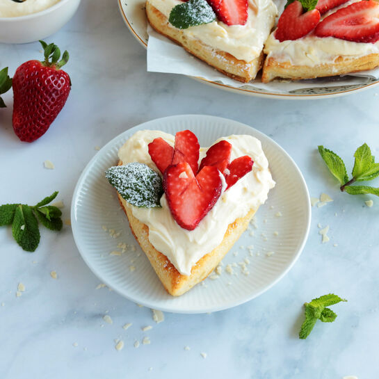 Herzförmiges Plundergebäck mit Frischkäsecreme und Erdbeeren auf kleinem Teller, dekoriert mit Minze
